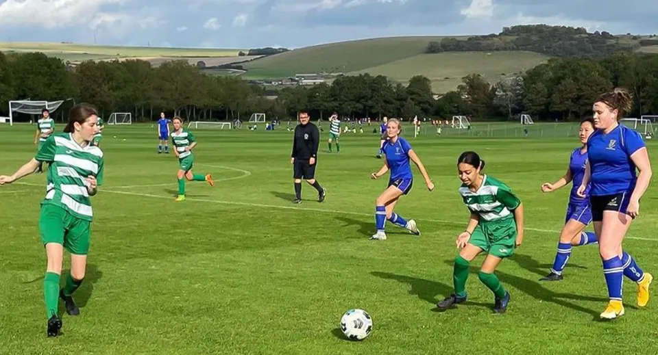 Studenti del Lancing College giocano una partita di calcio su un campo verde davanti a un paesaggio collinare e un cielo nuvoloso.