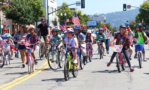Niños y adultos montan en bicicleta por una calle portando banderas estadounidenses durante un desfile