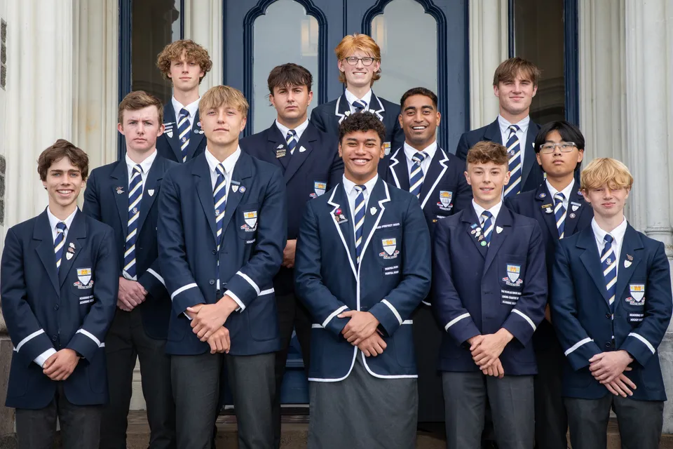 Un gruppo di studenti della Otago Boys' High School si trova davanti a un edificio con grandi finestre.