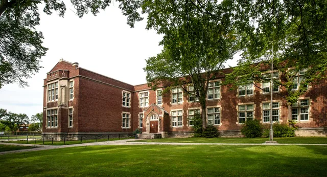 Das Backsteingebäude des Daniel McIntyre Collegiate Institute mit einem gepflegten Rasen und Bäumen im Vordergrund.