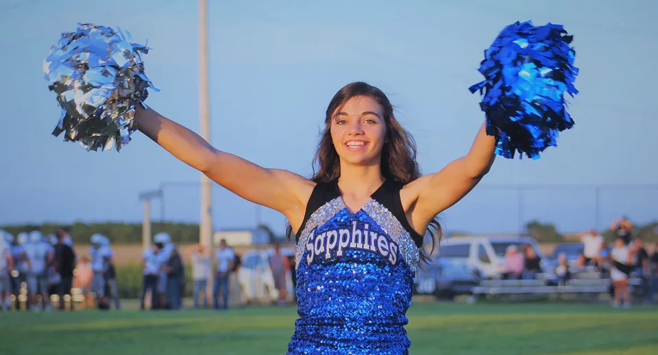Una cheerleader della Thunder Ridge High School è in piedi sul campo sportivo e agita pom-pom blu e argento.