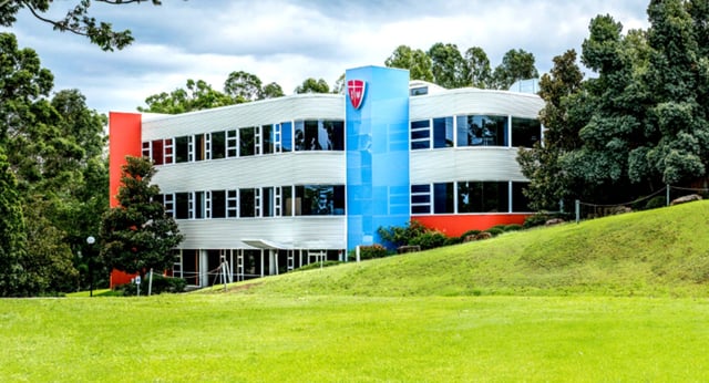 Das moderne Gebäude des John Paul College mit großen Fenstern steht auf einem grasbewachsenen Hügel, umgeben von Bäumen und Grünfläche.