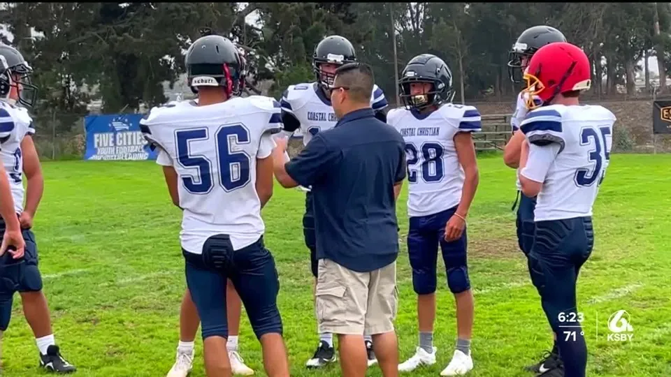 Giocatori di football americano della Coastal Christian School in divisa e casco intorno al loro allenatore sul campo.