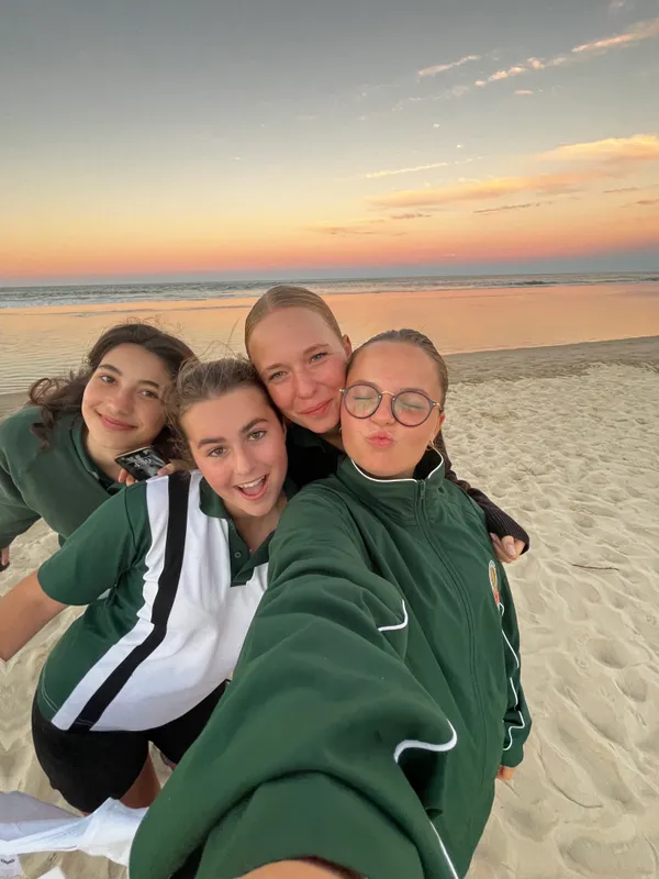 Selfie de cuatro alumnas con el uniforme verde del colegio en la playa al atardecer