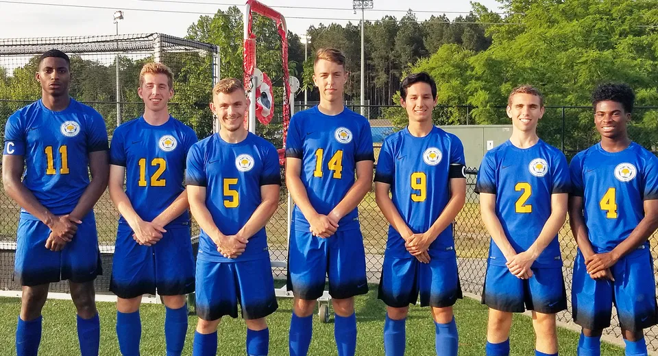 Un gruppo di giovani calciatori in maglia blu si trova sul campo sportivo della King George Secondary School.