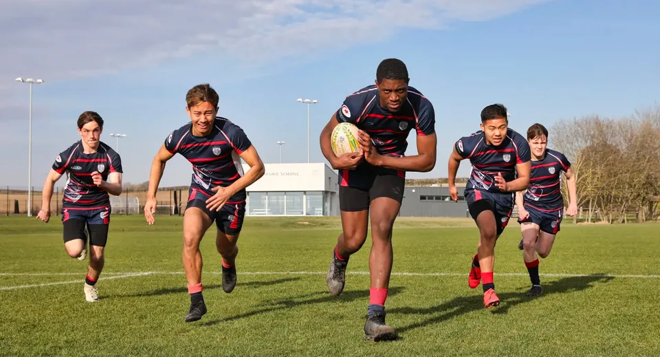 Studenti della Ashford School giocano a rugby su un campo d'erba davanti a un edificio sportivo.