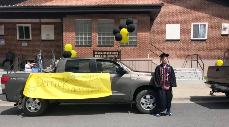 Un diplomato della Huntington High School in toga e tocco sta accanto a un pick-up decorato davanti all'edificio della scuola.