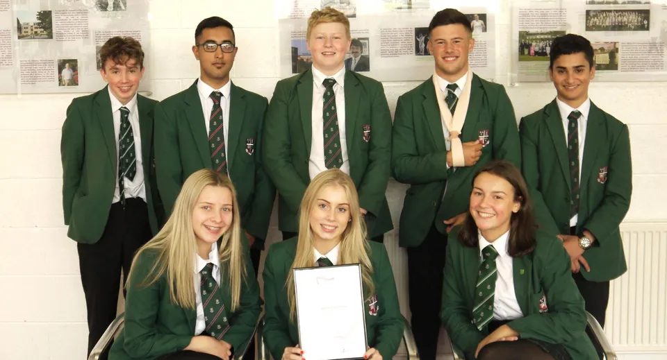 Un gruppo di studenti in uniforme verde posa per una foto di gruppo alla Woodhouse Grove School davanti a dei quadri sulla parete.