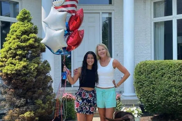 Chica joven sosteniendo globos y mujer rubia sonriendo frente a la entrada de una casa
