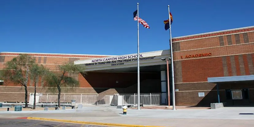Il grande edificio in mattoni della North Canyon High School è circondato da un'area pavimentata e da diverse bandiere americane nel cielo blu.