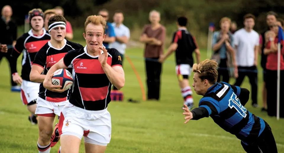 Studenti della King's Ely giocano una partita di rugby sul campo sportivo con spettatori sullo sfondo.