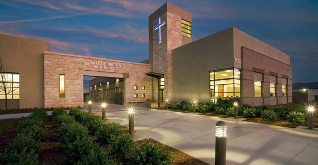Das mehrstöckige Gebäude der Valley Christian High School mit beleuchtetem Eingang und Türmchen vor einem bunten Abendhimmel.