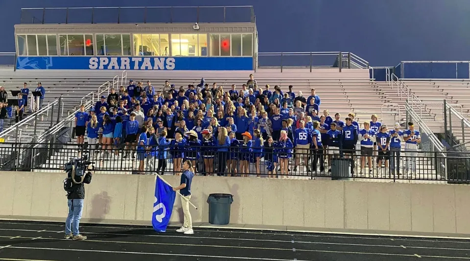 La squadra sportiva della Sparta High School si raduna sulle tribune di uno stadio con la scritta Spartans sullo sfondo.