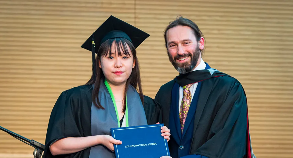 Una neodiplomata in toga sta in piedi insieme a un uomo in abito accademico e tiene in mano un diploma nel campus della ACS International School Cobham.