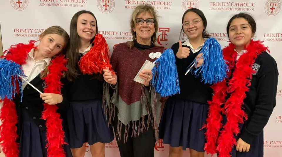 Un gruppo di studentesse con accessori di piume colorate sta davanti al logo della Trinity Catholic Jr/Sr High School.