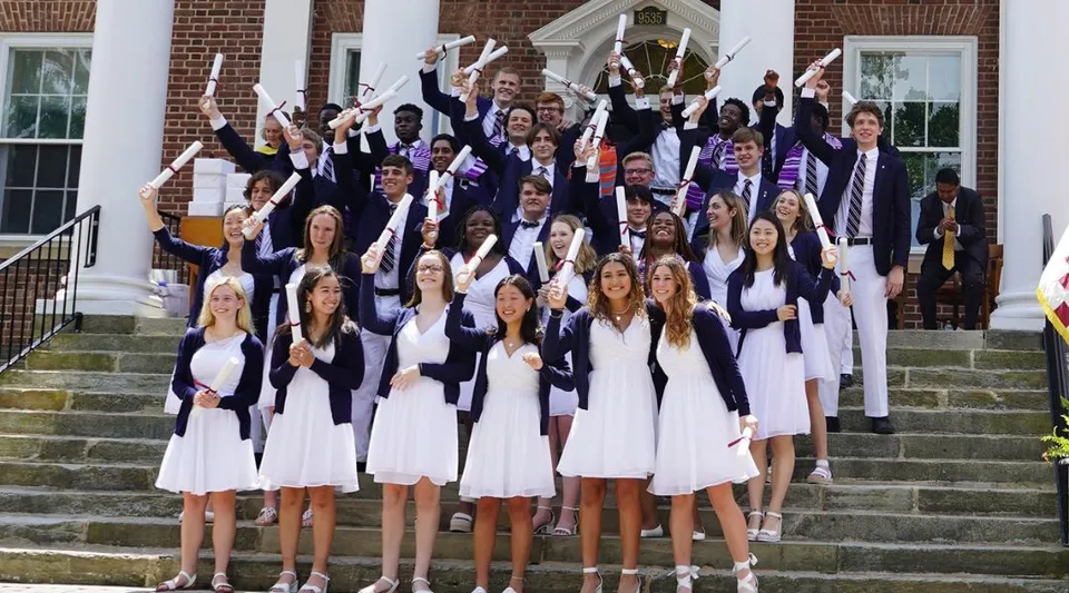 Un grande gruppo di diplomati è in piedi con toghe bianche e viola sui gradini di un edificio in mattoni della Saint James School.