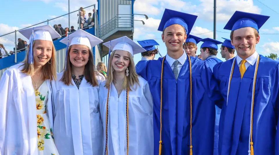 I diplomati della St. Mary's High School in toga blu sono davanti a un edificio scolastico sotto un cielo azzurro.