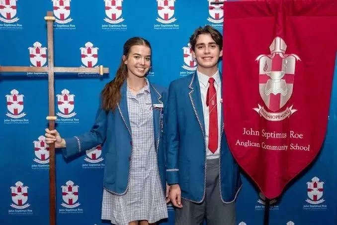 Dos estudiantes con el uniforme de la John Septimus Roe Anglican Community School posan con una cruz de madera y un estandarte rojo.