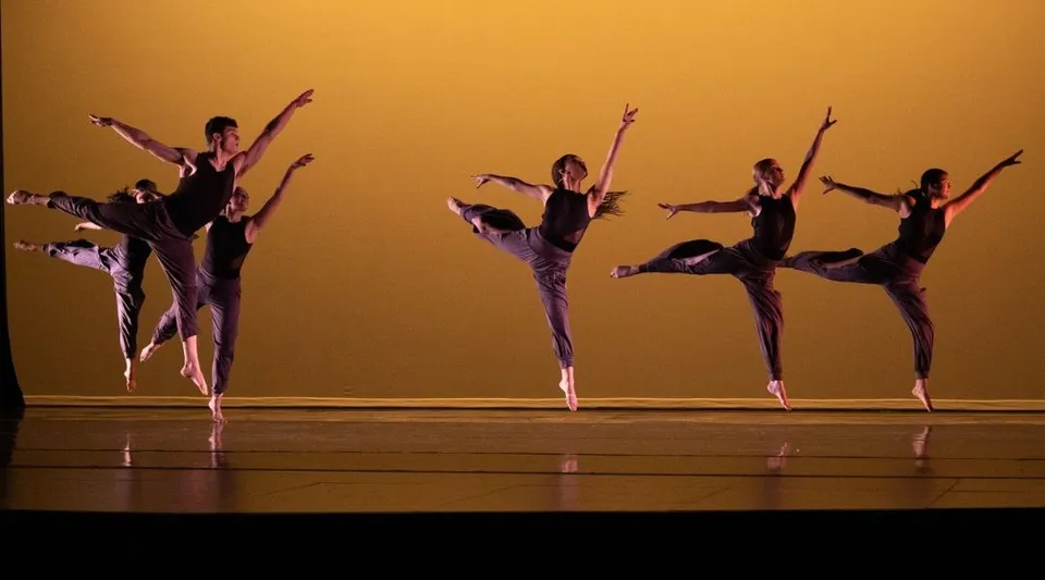 Un gruppo di ballerini e ballerine della Deerfield Academy si esibisce in una performance sincronizzata sul palco davanti a uno sfondo dorato.