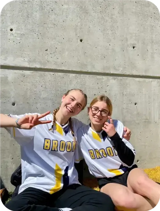 Due studentesse in maglia sportiva fanno il segno della pace sedute contro un muro