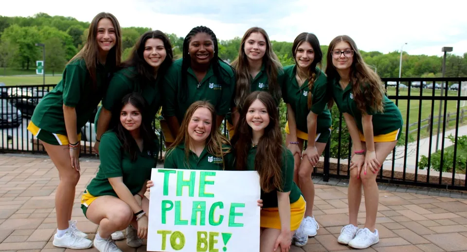 Un gruppo di studentesse della St. Mark High School con un cartello 'The Place to Be' davanti a un parco con alberi e recinzione.