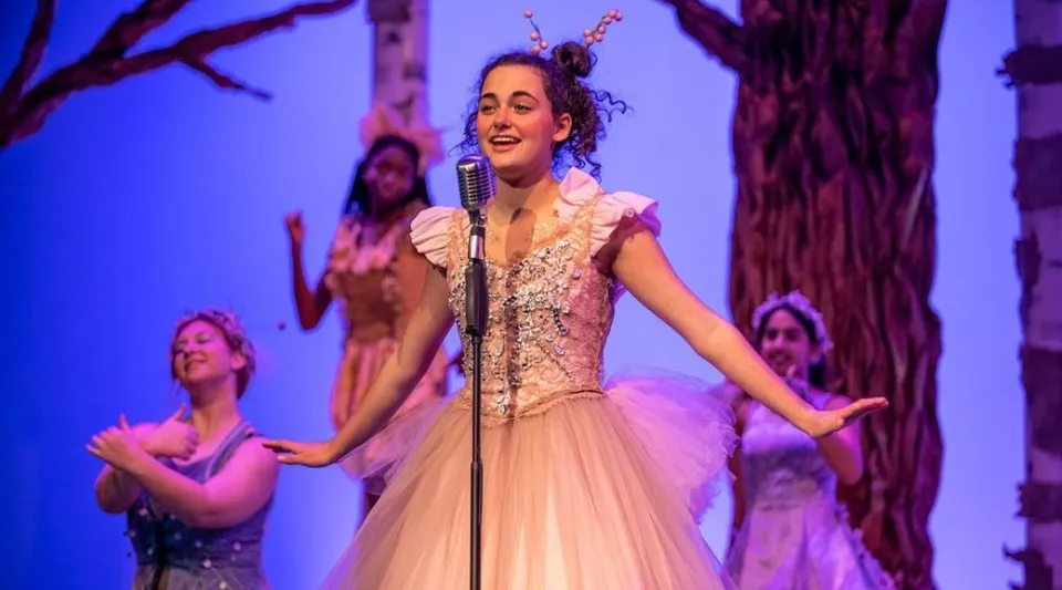 Una donna con un abito ornato di pizzo canta sul palco della Western Reserve Academy davanti a uno sfondo boschivo colorato, con danzatori sullo sfondo.