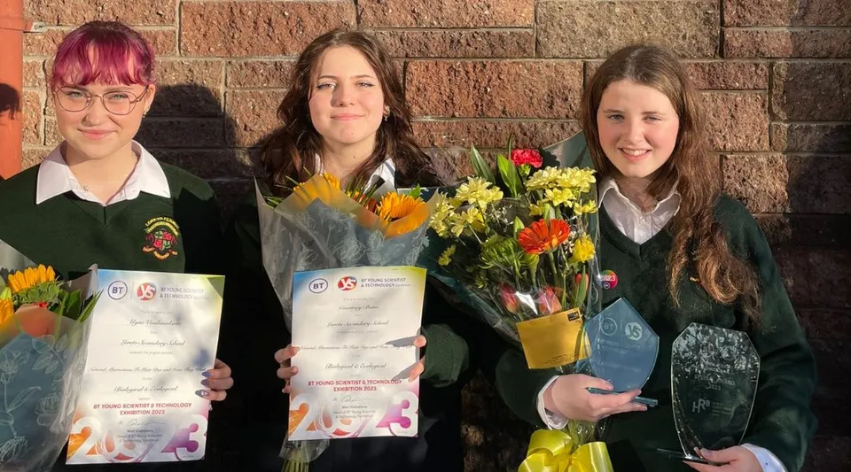 Tre studentesse della Loreto Secondary School a Cork tengono in mano certificati e fiori davanti a una parete in mattoni.