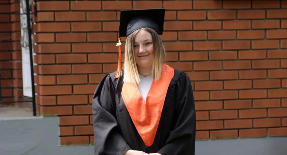 Una studentessa con la toga da diploma sta davanti a una parete di mattoni nel campus del Superior Collegiate & Vocational Institute.