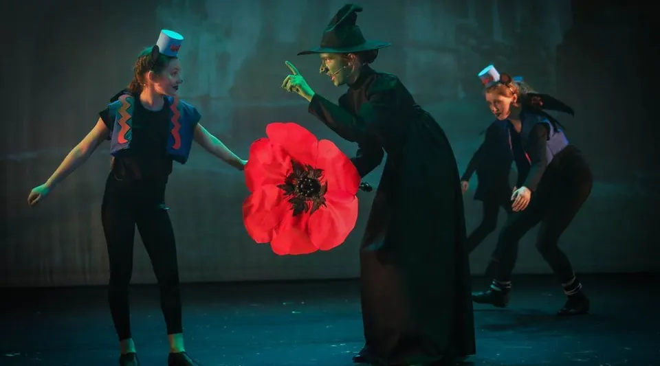 Spettacolo teatrale con studenti e studentesse della Howard of Effingham School, una persona indossa un mantello nero e un cappello, un grande papavero rosso è visibile in primo piano.