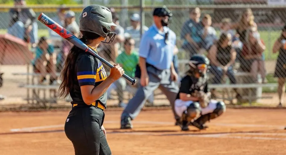 Una giocatrice di softball è in piedi sul campo di gioco del Mesa Unified School District, con altri giocatori e spettatori sullo sfondo.