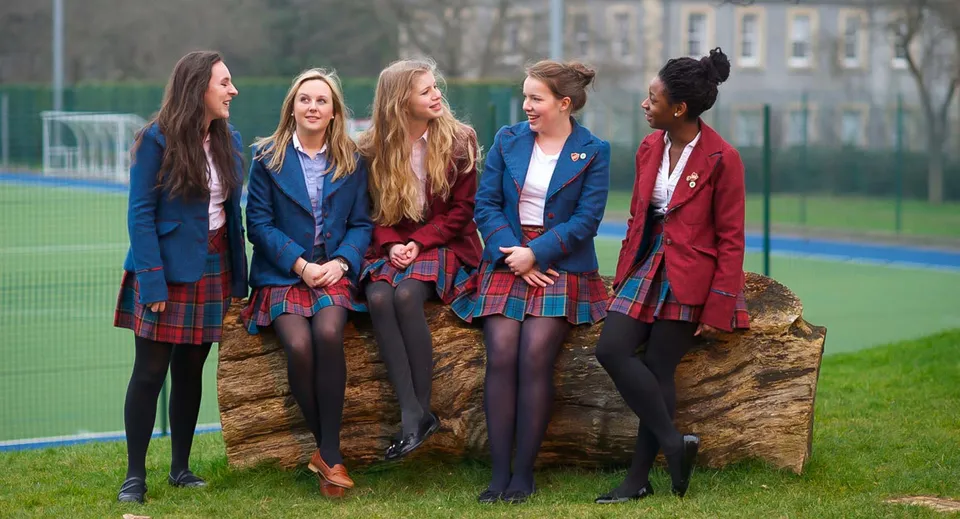 Un gruppo di studentesse della St. George's School in divisa scolastica si trova insieme sul campo sportivo davanti agli edifici.