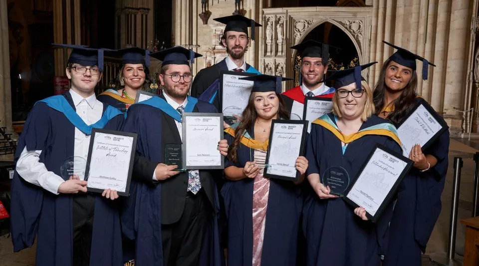 Un gruppo di diplomati del York College si trova insieme in abito accademico in uno splendido interno e tiene in mano i propri diplomi.