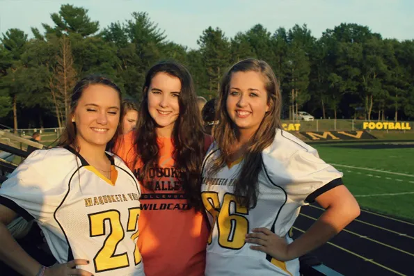 Tres jóvenes sonrientes con camisetas deportivas posan en un campo de fútbol americano