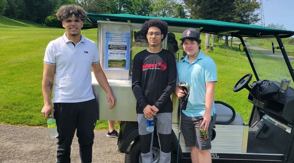 Tre studenti della Harmony Christian School sono davanti a una golf cart su un prato con alberi sullo sfondo.