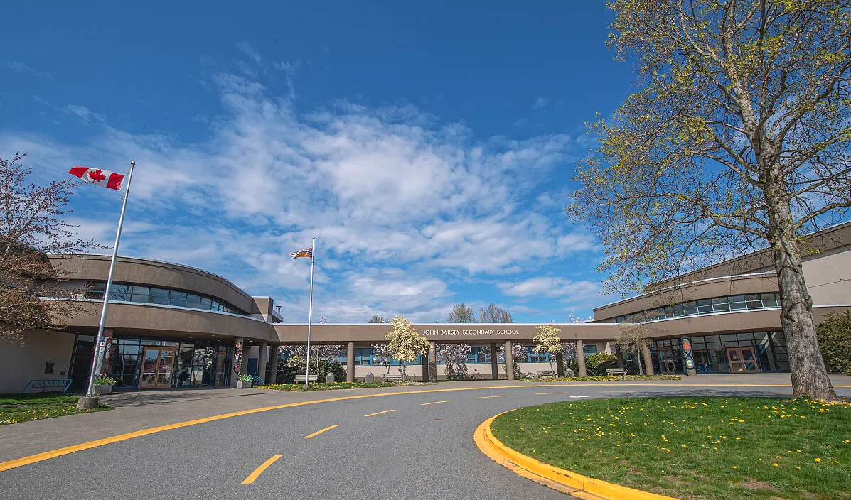 El edificio moderno de la John Barsby Secondary School está rodeado de árboles y muestra una bandera canadiense ante un cielo azul.