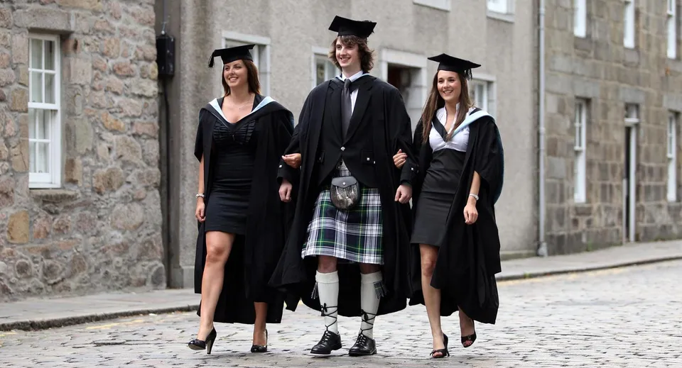 Tre diplomati del Fettes College in abito accademico sono in piedi davanti a un edificio in pietra su una pavimentazione in ciottoli.