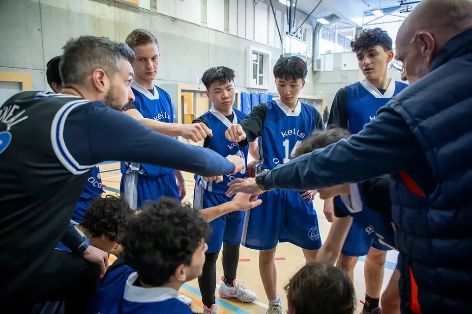 Un gruppo di giovani giocatori di basket in maglia blu ascolta le istruzioni dell'allenatore nell'area della Kells Academy.