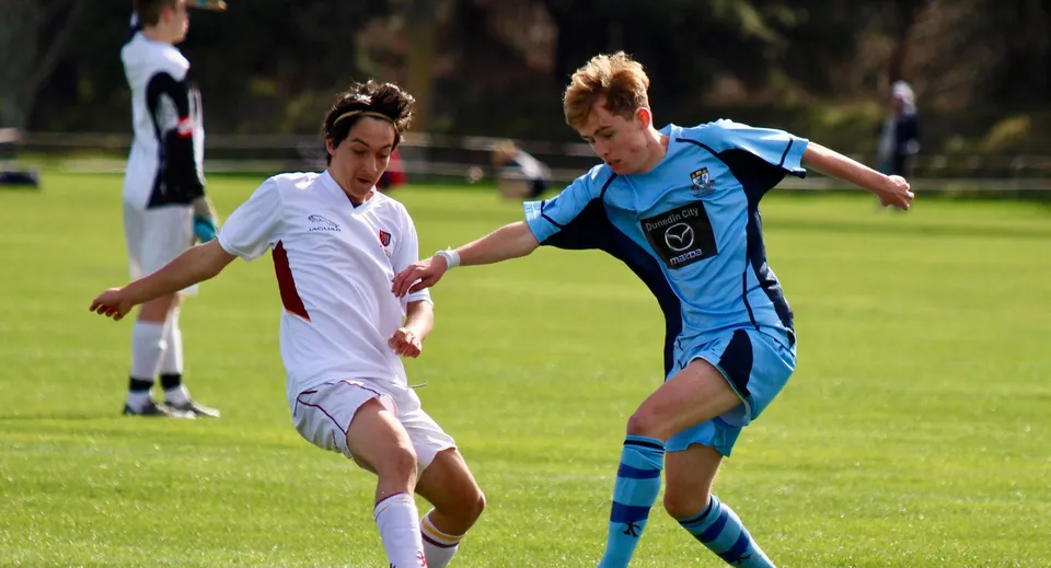 Due studenti della King's High School giocano a calcio su un campo con altri giocatori sullo sfondo.