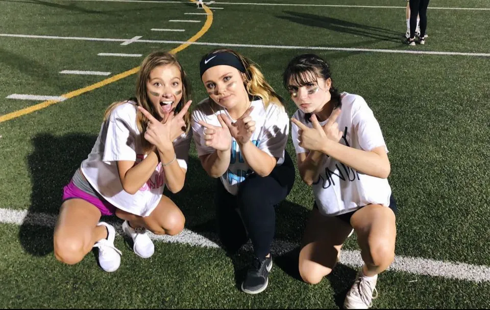 Tre studentesse della Monarch High School posano sul campo sportivo con le linee di gioco e le porte sullo sfondo.