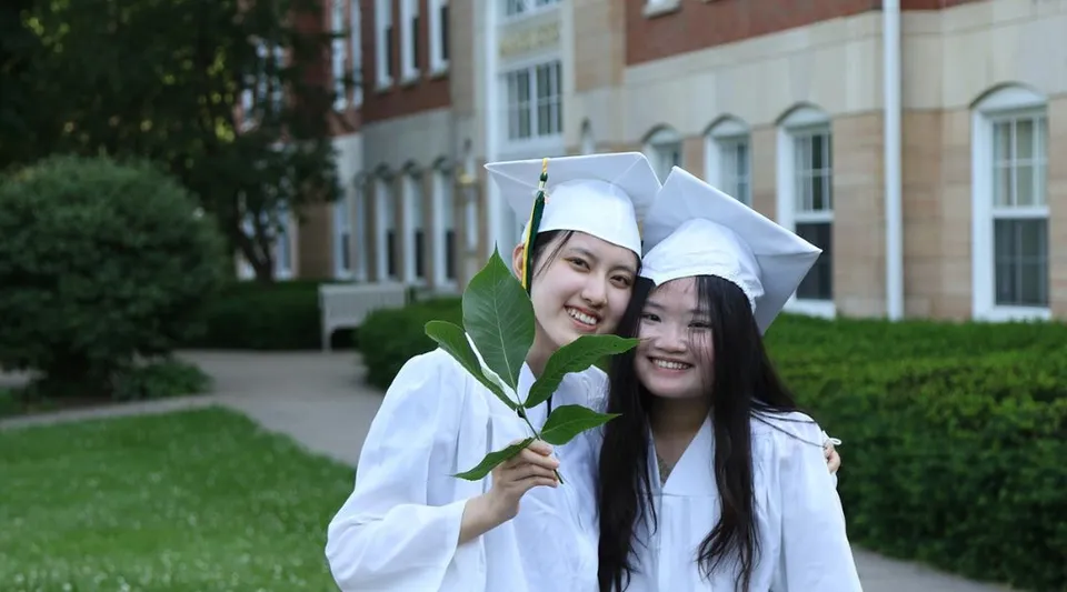 Due studentesse in toga da diploma tengono insieme una foglia verde nell'area della Maharishi School.
