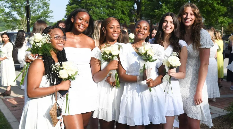 Un gruppo di studentesse della Shady Side Academy in abiti bianchi e con mazzi di fiori si trova in un giardino con alberi e spazi verdi.