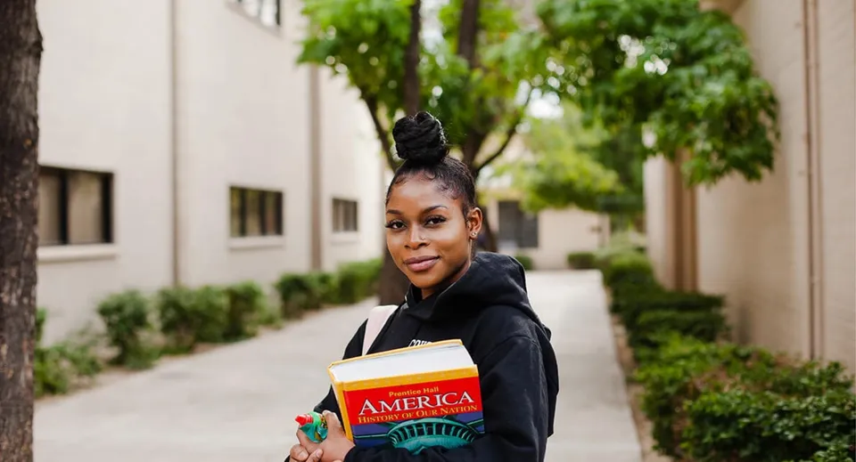 Una giovane donna con un'acconciatura vistosa cammina lungo una strada alberata portando un libro nel campus della Phoenix Christian Preparatory School.