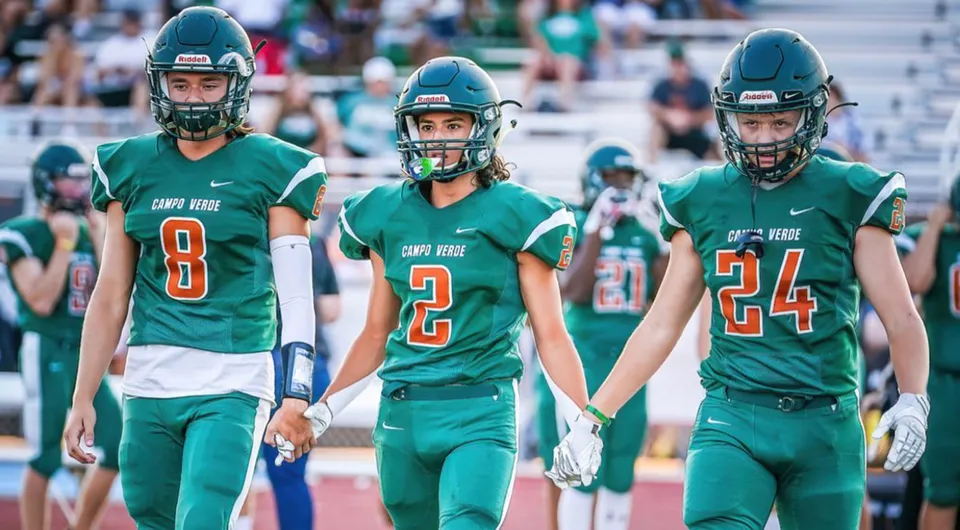 Tre giocatori di football della Campo Verde High School in maglia verde sono sul campo da gioco davanti agli spettatori.