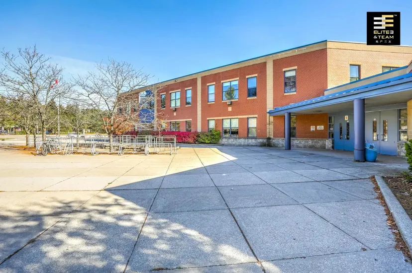 L'edificio in mattoni della Oakville Trafalgar High School con dettagli blu e rossi si trova dietro un cortile pavimentato e alberi.
