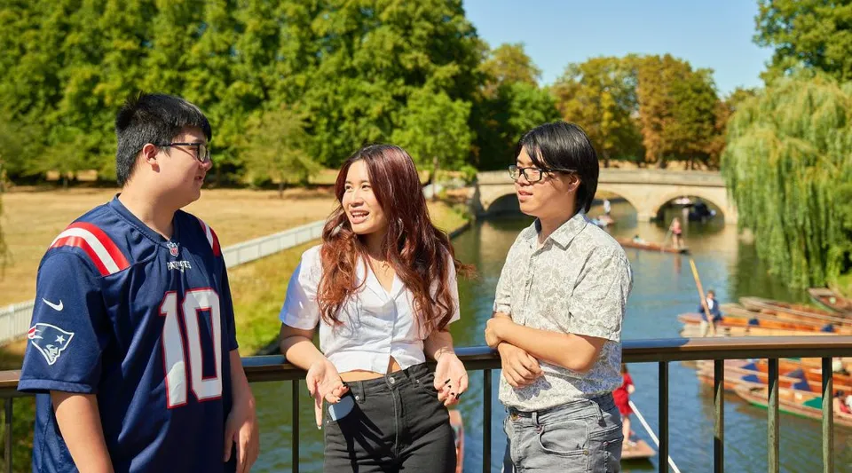 Tre studenti della CATS Cambridge sono insieme e chiacchierano in un'area tipo parco con alberi e acqua.