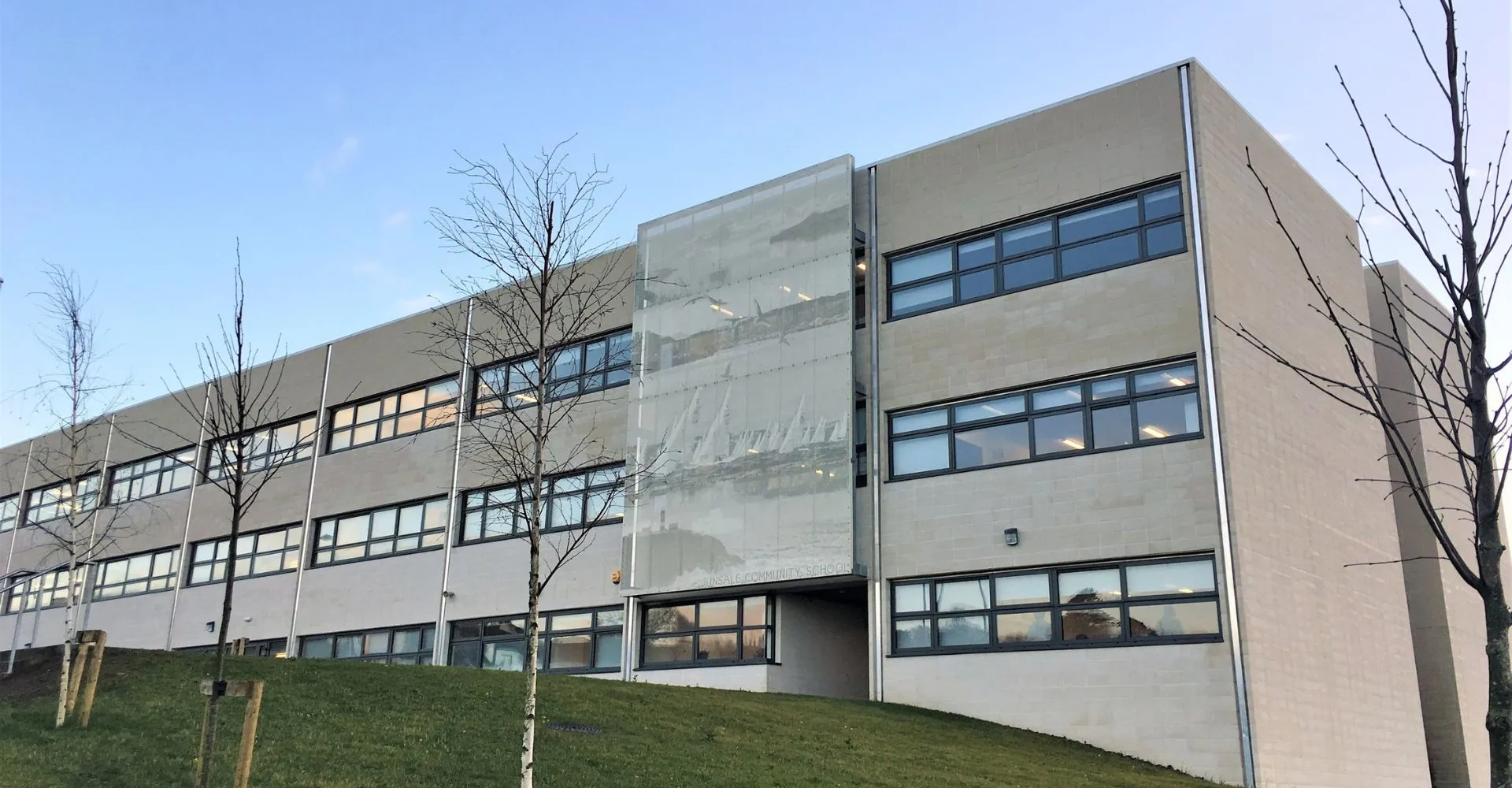 L'edificio moderno a più piani della Kinsale Community School si trova su un pendio erboso con alberi spogli sullo sfondo.