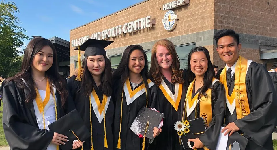 Un gruppo di diplomati in toga è in piedi davanti al Mclean Sports Centre sul campus della Burnaby South School.