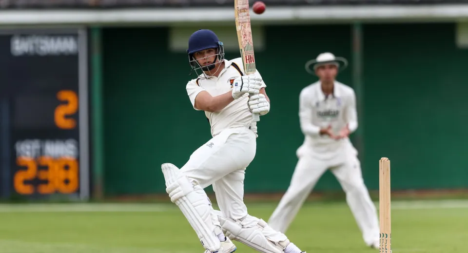 Un giocatore di cricket in divisa bianca si trova sulla battuta mentre un altro giocatore osserva sul campo da gioco del Bishop’s Stortford College.