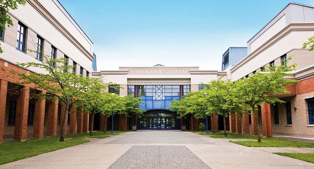 El edificio de ladrillo de la Burnaby South School con grandes ventanas y un cartel se encuentra al final de un camino pavimentado entre plantas verdes.