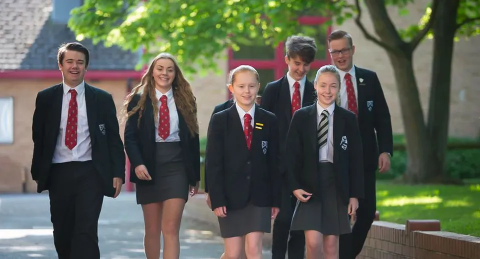 Un gruppo di studenti e studentesse in divisa scolastica cammina insieme nel campus della Fulston Manor con alberi ed edifici sullo sfondo.
